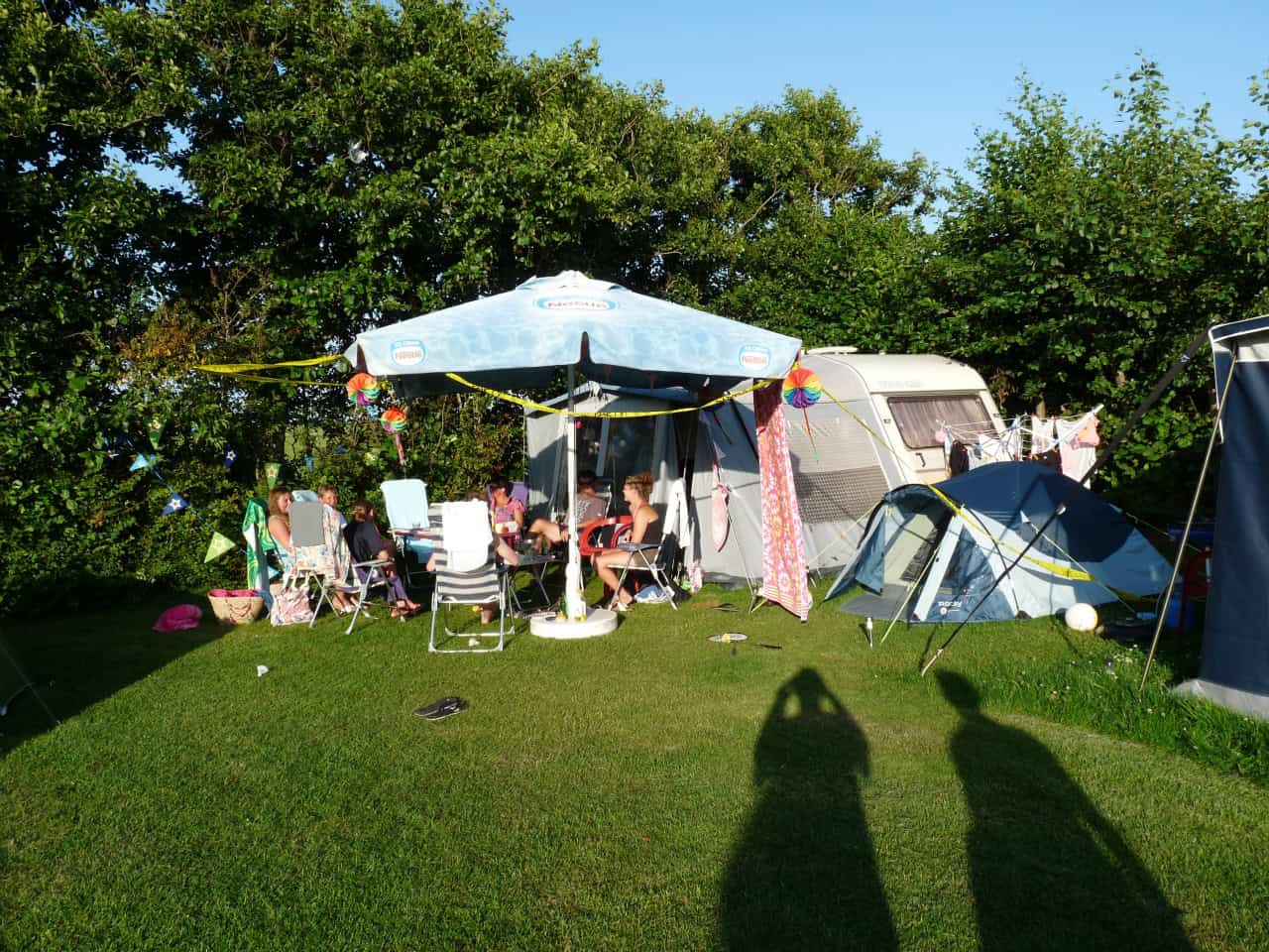 Gasten onder een parasol bij caravan en tent in de avondzon